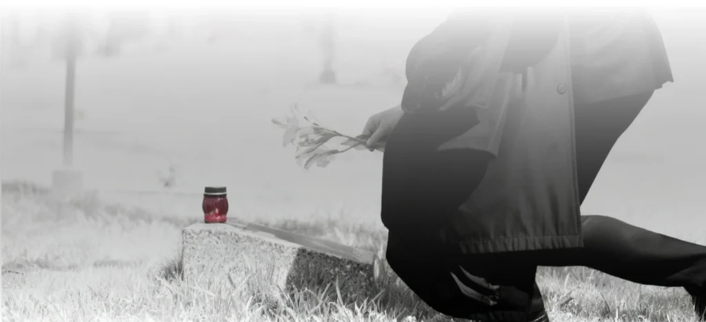 A person dressed in dark clothing kneels at a gravesite, holding a small bouquet of flowers. A red memorial candle sits on the gravestone. The surrounding scene is muted and foggy, emphasizing a somber mood.