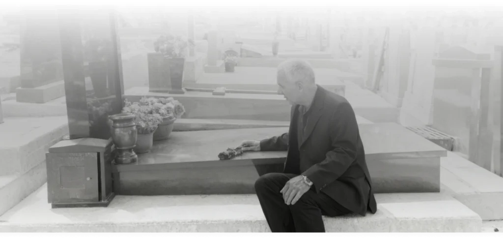 Person sitting by a tombstone in a graveyard.
