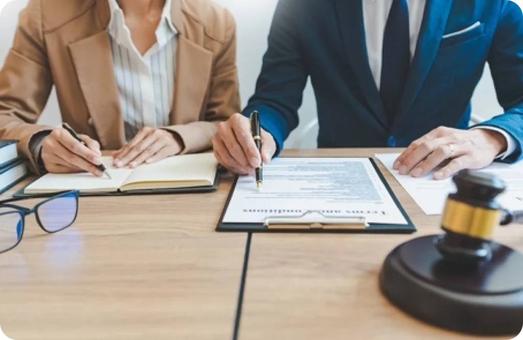 Two professionals in formal attire reviewing and signing legal documents at a desk with a gavel, notebook, and eyeglasses.