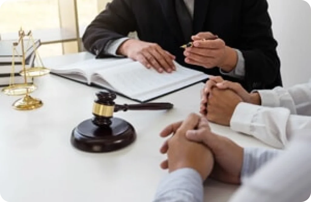 Image of a legal consultation scene featuring a gavel and scales of justice on a table. Two individuals are seated across from a person who appears to be a lawyer or legal advisor, engaged in discussion with an open book in front of them.