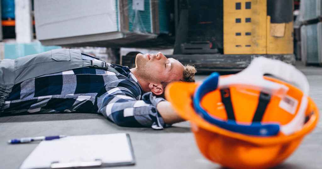 An unconscious worker lies on a warehouse floor next to a fallen hard hat, illustrating a workplace injury.