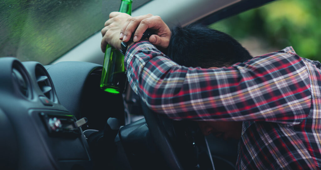 Drunk man slumped over a car steering wheel holding a bottle.