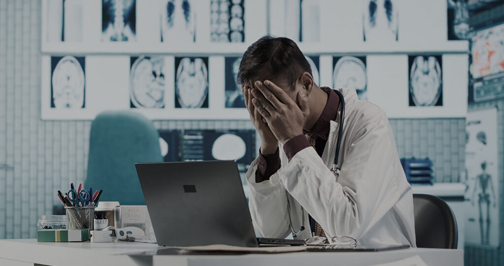 Stressed doctor covering his face at a desk with laptop.