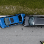 Aerial view of a blue car and black SUV after a crash on a road, with a responder standing nearby.