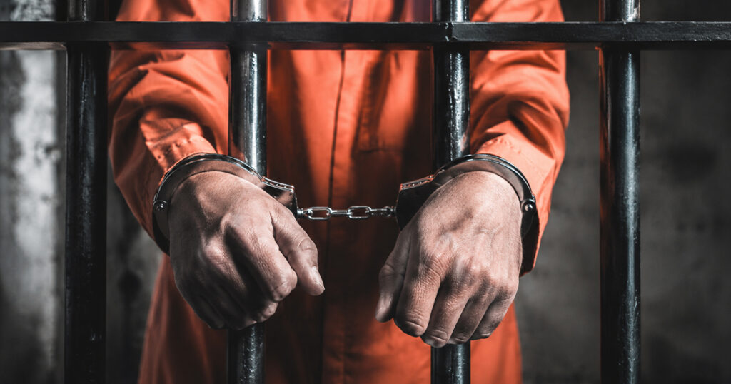 Prisoner in an orange jumpsuit stands behind jail bars with hands cuffed in front.