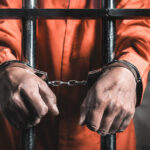 Prisoner in an orange jumpsuit stands behind jail bars with hands cuffed in front.