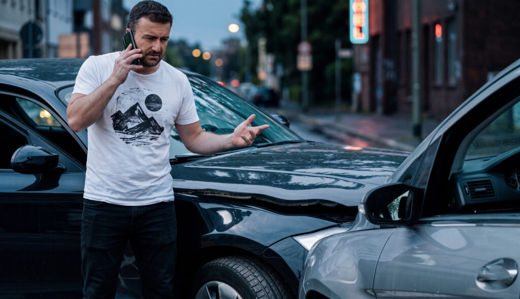 Man standing by his damaged car on a city street, talking on his phone with concern.