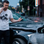 Man standing by his damaged car on a city street, talking on his phone with concern.
