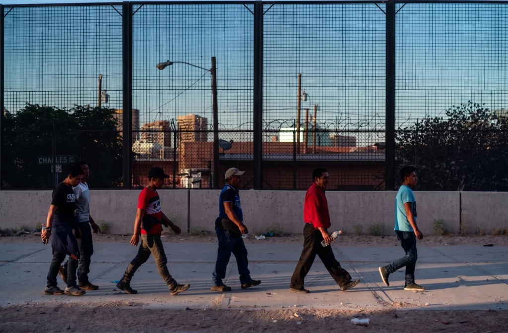 Men walking along a sidewalk next to a tall mesh border fence.