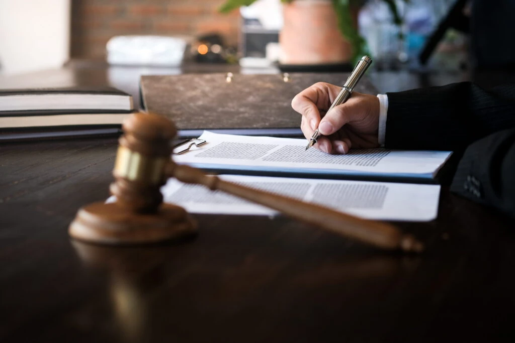 Close-up of an attorney signing contract papers with a fountain pen at a dark wooden desk featuring a judge's gavel.