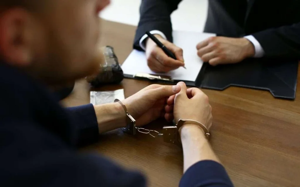 Handcuffed suspect being questioned by an official taking notes at a desk.