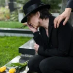 Mourning woman kneeling by gravestone comforted by supportive hand.