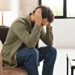 Stressed man sitting on sofa holding head near laptop computer
