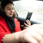 A woman in a red coat looking at her smartphone while holding the steering wheel of a car.